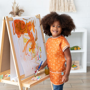 A child with curly hair joyfully paints vibrant brushstrokes on an easel, wearing a Bumkins Short-Sleeved Smock: Grounded.