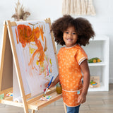 A child with curly hair joyfully paints vibrant brushstrokes on an easel, wearing a Bumkins Short-Sleeved Smock: Grounded.