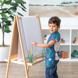 A young boy in a Bumkins Short-Sleeved Smock: Animal Prints paints on an easel in a bright room with a large plant and shelves as a backdrop.