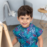 A child beams at the camera in a Bumkins Short-Sleeved Smock: Animal Prints, beside an easel, gray chair, and wooden table.