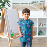 A child in a Bumkins Short-Sleeved Smock: Animal Prints grins next to an easel amid shelves of toys in a bright room.