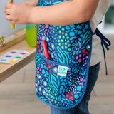 Close-up of a child in a Bumkins Short-Sleeved Smock: Animal Prints next to an easel with a red paintbrush in the pocket.