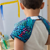 A child in a Bumkins Short-Sleeved Smock: Animal Prints stands by an easel with brushes nearby, with the hook-and-loop neck closure visible.