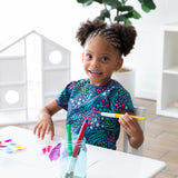 A child paints happily at a white table, wearing a Bumkins Short-Sleeved Smock: Animal Prints in a bright, airy room.