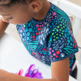 A child paints on paper in while wearing a Bumkins Short-Sleeved Smock: Animal Prints.
