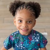 A smiling girl with braided hair in a Bumkins Short-Sleeved Smock: Animal Prints sits on a chair against a wooden floor.