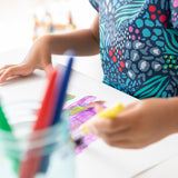 Close-up of a child in a Bumkins Short-Sleeved Smock: Animal Prints painting on paper; a jar of brushes is in the foreground.