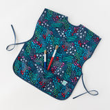 Bumkins Short-Sleeved Smock: Animal Prints lays on a white background, holding two paintbrushes in the pocket.