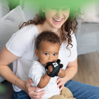 A woman smiles as her baby enjoys a Bumkins Silicone Un-Droppable Mitt Teether: Mickey Mouse on the couch, surrounded by green leaves.