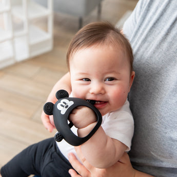 A baby chews on a Bumkins Silicone Un-Droppable Mitt Teether: Mickey Mouse, cradled by an adult.