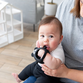 A baby sits on an adult's lap indoors, nibbling a Bumkins Silicone Un-Droppable Mitt Teether: Mickey Mouse.