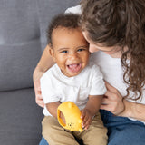 A smiling baby sits on a woman's lap on a gray couch, holding a Bumkins Silicone Un-Droppable Mitt Teether: Winnie the Pooh.