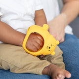 A child holds a Bumkins Silicone Un-Droppable Mitt Teether: Winnie the Pooh, while sitting on an adult’s lap.