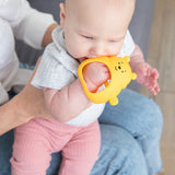 A baby with grey top and pink pants chews on a Bumkins Silicone Un-Droppable Mitt Teether: Winnie the Pooh while on an adults lap.