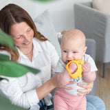 A smiling woman holds a baby with a Bumkins Silicone Un-Droppable Mitt Teether: Winnie the Pooh in a cozy room, plant in view.