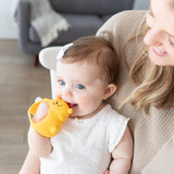 A baby with blue eyes chews a Bumkins Silicone Un-Droppable Mitt Teether: Winnie the Pooh, while held by a smiling woman in a beige sweater.