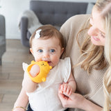 A baby with a white bow chews on the Bumkins Silicone Un-Droppable Mitt Teether: Winnie the Pooh while sitting on a woman's lap.