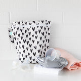 A Bumkins Wet Bag 2 Pack: Hearts and Rainbows sits on a white table against a white wall, with cloth diapers and breast pump parts in front.
