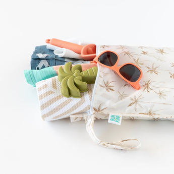Bumkins Wet Bag: Palm Daze holds sunglasses, a toy palm tree, and beach items against a white background.
