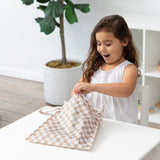 A young girl peers into a Roamio by Bumkins Wet Bag: Sand Check at a white table, with a plant in the background.