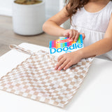 A child puts a Bumkins Wet Bag 2 Pack: Sand and Black Check into a beige checkered diaper bag on a white table.