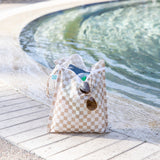 Bumkins Wet Bag + Wet/Dry Bag Set in Sand Check sits poolside as sunlight sparkles on the water.