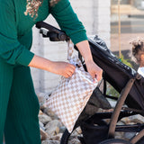 An adult strolls outdoors with a child in a stroller and Bumkins Wet Bag 2 Pack: Sand and Black Check attached to the handle.