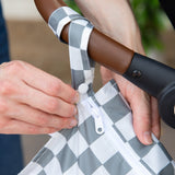 Close-up of a person attaching a Bumkins Wet Bag: Charcoal Check to a stroller handle using the snap button on the handle.