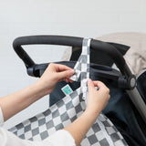 Hands attaching a Bumkins Wet Bag: Charcoal Check to a stroller handle.