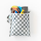 A bag from Bumkins Wet Bag + Wet/Dry Clutch Set: Charcoal Check holds flip-flops, a towel, and a yellow toy dinosaur on a white background.