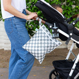 A person wearing jeans and a white shirt pushes a child in a stroller outdoors, with a Bumkins Wet Bag: Charcoal Check attached to the handle.