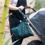 A person in black overalls pushing a stroller outdoors with a Bumkins Wet Bag: Starry Cactus attached to the handle.