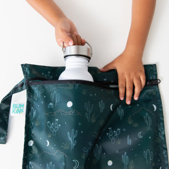 A child places a white water bottle into a Bumkins Wet Bag: Starry Cactus on a white table.