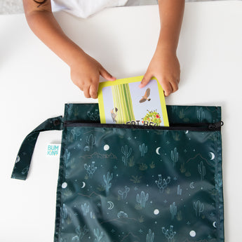 A child slips a book into a Bumkins Wet Bag: Starry Cactus, featuring cactus and moon designs, on a white table.
