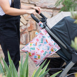Someone attaches the Bumkins Wet Bag + Wet/Dry Bag Set: Boots, an eco-friendly bag, to a stroller outdoors near plants.