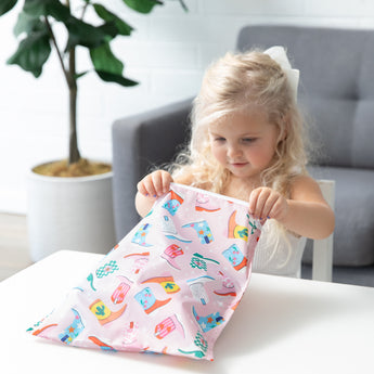 A young girl opens a bag from Bumkins The Playdate Planner Deluxe 5-Piece Set: Boots at a white table.