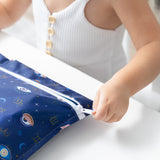 A child in a white top unzips a Bumkins Wet Bag: Evil Eye at a white table.