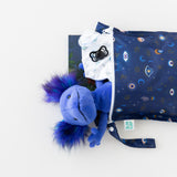 A plush blue toy with purple hair peeks out of the Bumkins Evil Eye Wet/Dry Bag, a cosmic book tucked inside.