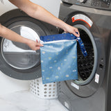 A person holds a Bumkins Wet Bag: Evil Eye in front of an open washing machine, with a white laundry basket in the background.