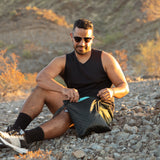 A man in sunglasses reaches into a Bumkins Wet Bag 2 Pack: Sand and Black Check while sitting on rocky ground in golden sunlight.
