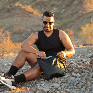 A man in sunglasses reaches into a bag from Bumkins The Chaos Coordinator Deluxe 5-Piece Set: Rich Black while sitting on rocky ground.