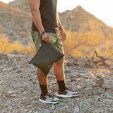 A man stands on rocky ground holding a Roamio by Bumkins Wet Bag: Rich Black with mountains in the background.