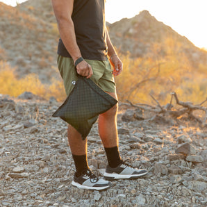 A man holds a bag from Bumkins Wet Bag + Wet/Dry Clutch Set: Rich Black while standing on rocky ground with mountains in the background.