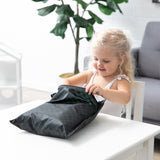 A child eagerly opens a Bumkins Wet Bag: Rich Black at a white table, with a plant and couch nearby.