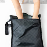 A child’s hands reach into a Bumkins Wet Bag 2 Pack in Sand and Black Check, made of waterproof fabric on a white surface.