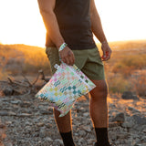 A person wearing green shorts and a black shirt stands on rocky ground at sunset, holding a Roamio by Bumkins Wet Bag: Motley Check.