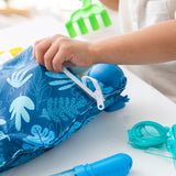 Close-up of a child's hand opening a Bumkins Wet Bag: Blue Tropic amid pool toys on a white table