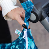 Close-up of a hand with black nails attaching a Bumkins Wet Bag: Blue Tropic to a stroller handle using the snap button on the strap.