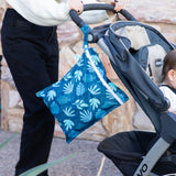 A person pushes a stroller with a child outdoors, with a Bumkins Wet Bag: Blue Tropic attached to the handle.
