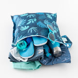 Bumkins Wet Bag: Blue Tropic holds a swimsuit, sun hat, goggles, water shoes, sunglasses, and a dinosaur toy on a white background.